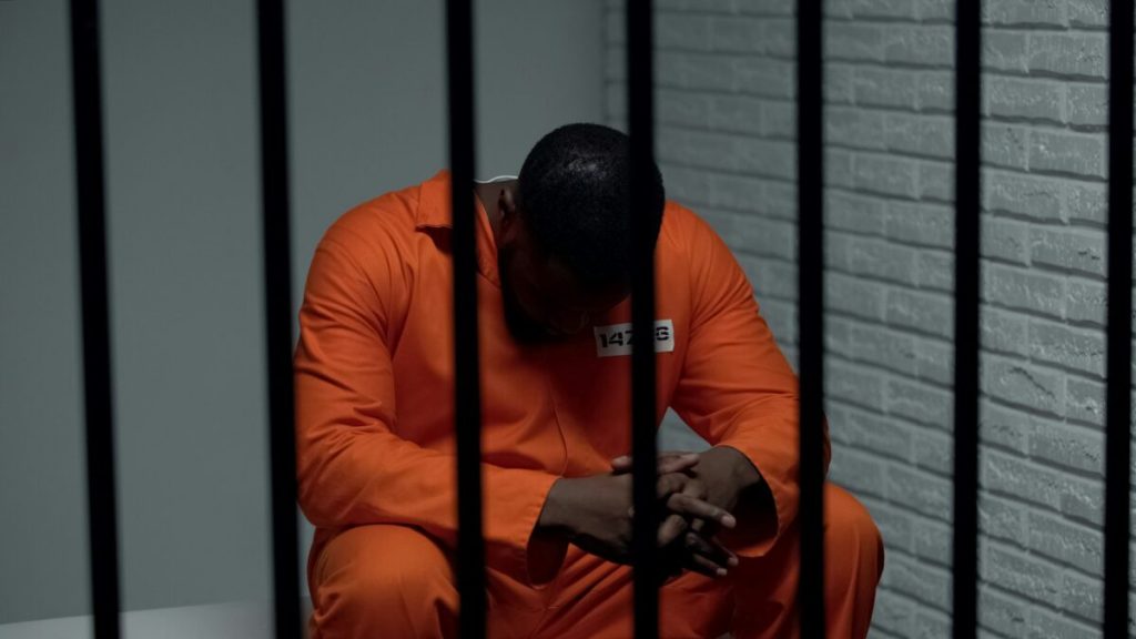 A man sits behind jail bars, head bowed.