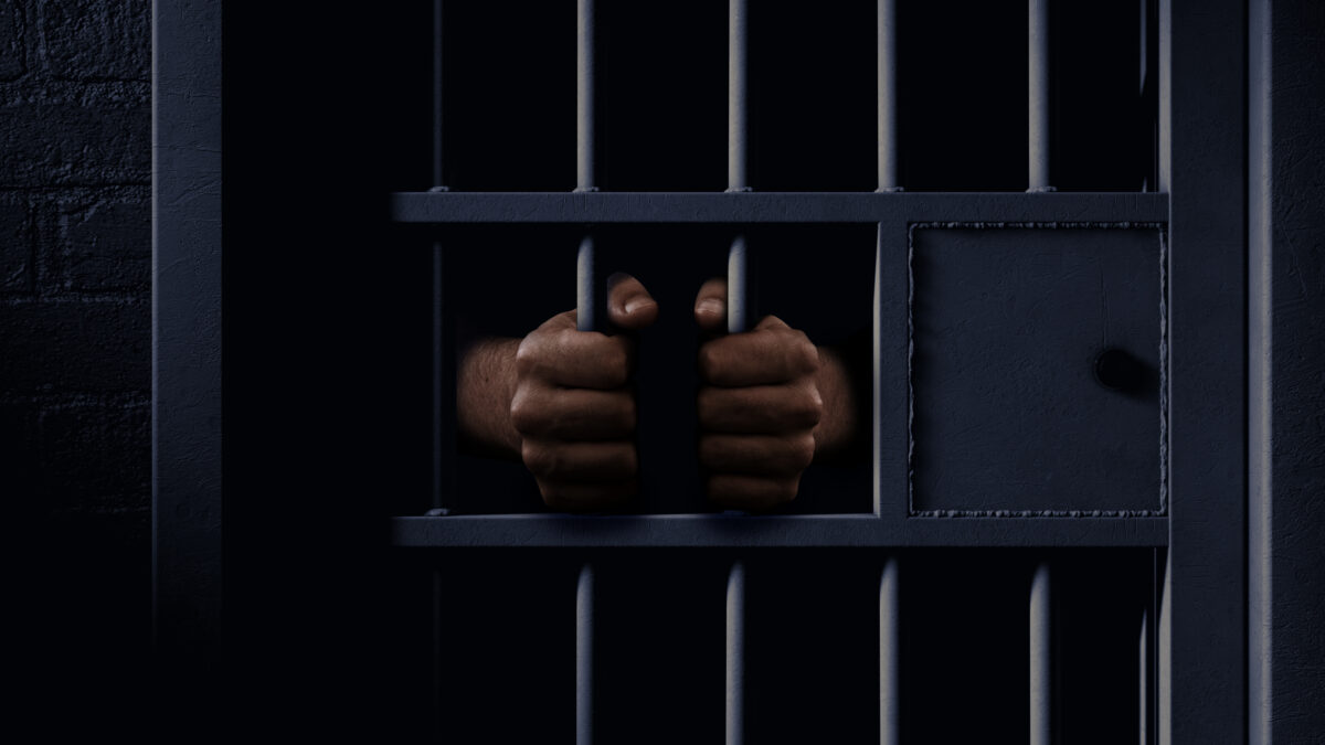 A closeup of a dimly lit prison holding cell door with a pair of african black hands holding onto the bars