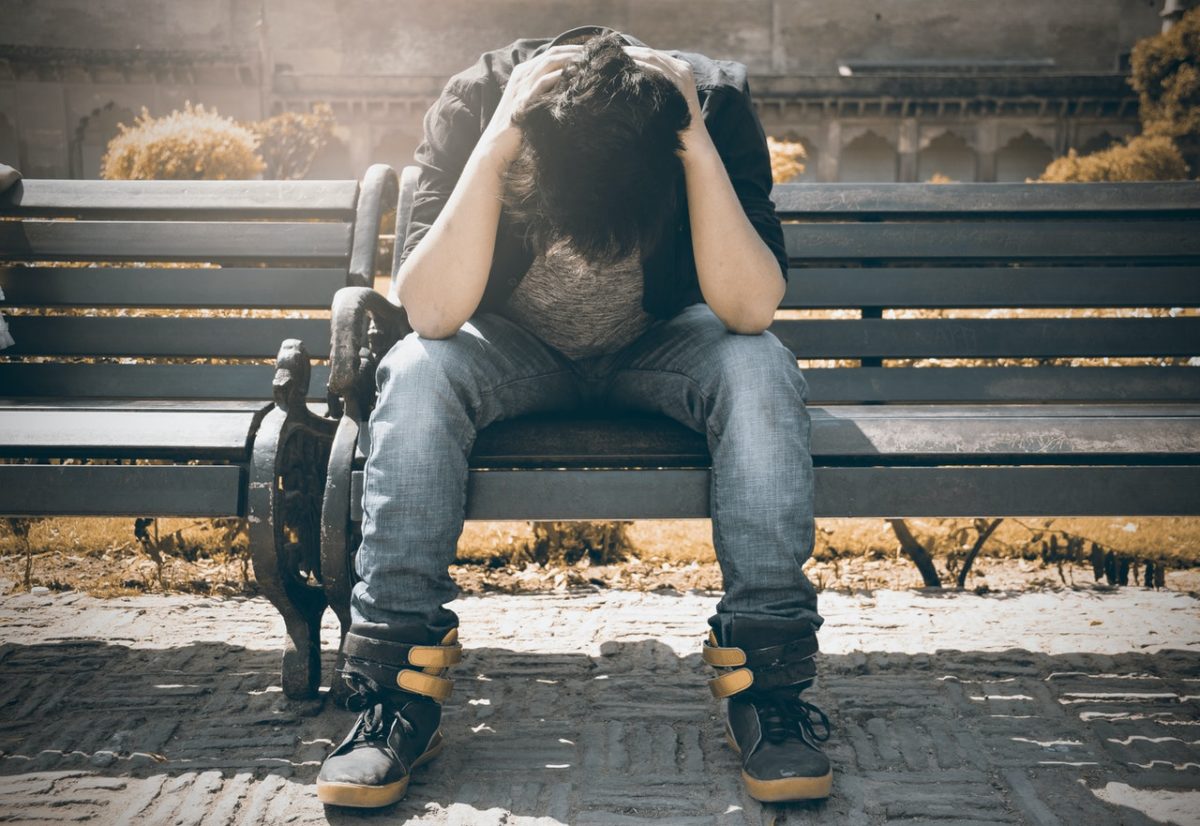 A young man is sitting on a park bench, looking worried.